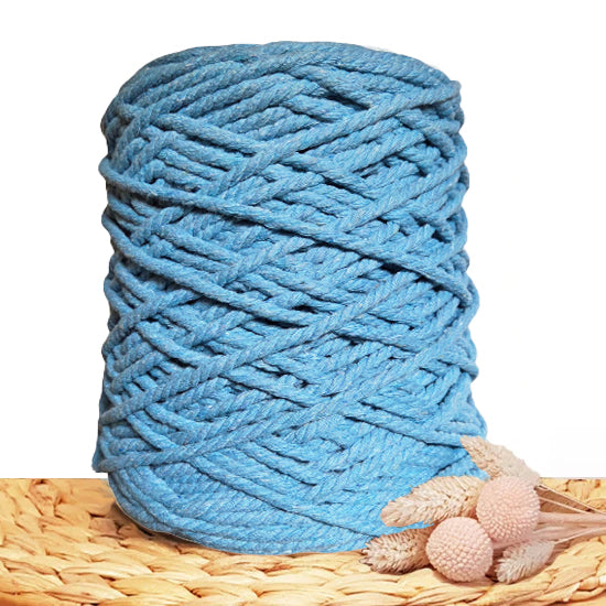 A large spool of thick 3ply cotton cord in a dusty light blue color, styled on a woven straw mat with dried pink flowers against a white background.