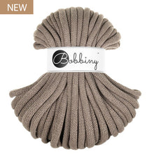 A skein of chunky braided cord in a coffee or taupe, wrapped with a white paper band featuring the 'Bobbiny' brand logo. A small brown 'NEW' label is in the top-left corner.