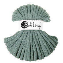 A skein of chunky braided cord in a muted sage green color, wrapped with a white paper label featuring the brand name 'Bobbiny' in black script and logo.