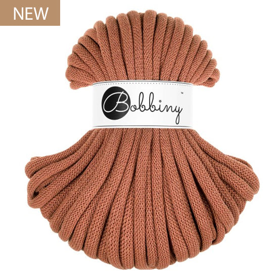 A skein of chunky braided cord  in a warm terracotta shade, wrapped with a white paper label that reads 'Bobbiny'. A small 'NEW' badge is visible in the top-left corner.
