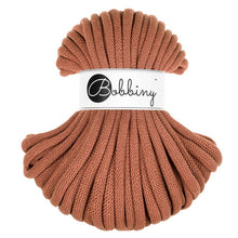 A skein of chunky braided cord in a warm terracotta or rust color, wrapped with a white paper label that reads 'Bobbiny' in black script and logo, set against a plain white background.